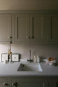 A stylish, mushroom-coloured Shaker galley kitchen features a cooker and sink run with Silestone worktop, wall cupboards and a single sink with a silver tap. Beautifully decorated with simple, everyday kitchen items such as muted ceramics, framed illustrations, a plastic bag full of bread, and small cleaning equipment, this calming and functional kitchen space blends character with peace.