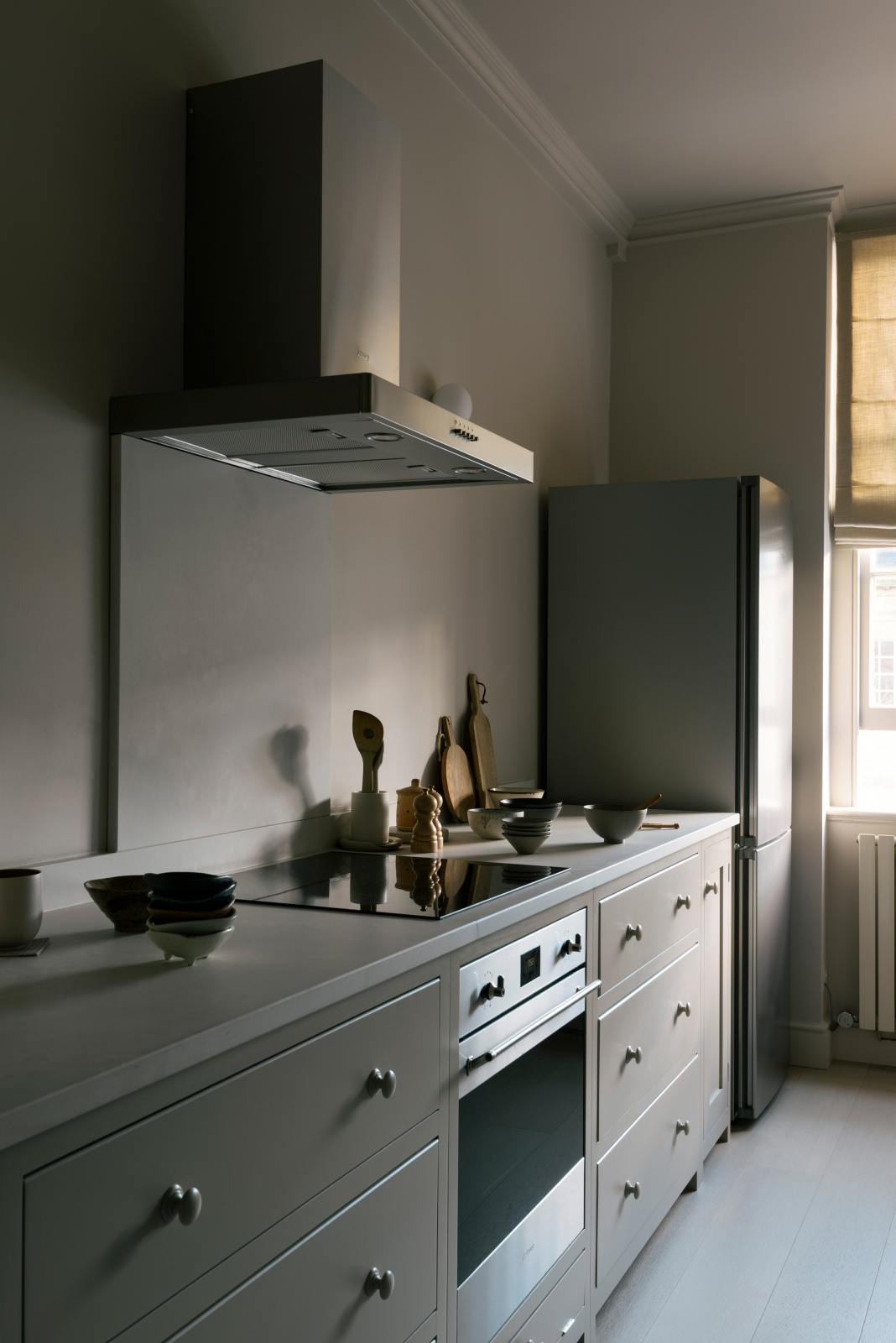 An angled view of a stylish, mushroom-coloured Shaker galley kitchen features a cooker and sink run with Silestone worktop. Beautifully decorated with simple, everyday kitchen items such as muted ceramics, wooden salt and pepper grinders, chopping boards, and an electric hob, this calming and functional kitchen space blends character with peace.