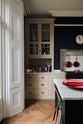 A quiet corner of a simple and elegant mushroom-brown Shaker kitchen features a cooker run with freestanding cupboards, a Carrara marble worktop, a false chimney breast, white porcelain splashback tiles, and a large kitchen island. The space feels loving and functional, adorned with artisan ceramics, copper pans, bright red rhubarb, luscious houseplants, and books.