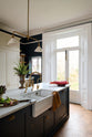 An angled view of a simple and elegant mushroom-brown Shaker kitchen features a large kitchen island with a single Belfast sink, a small section of copper worktop, and a bank of fitted cupboards housing a fridge-freezer and storage pantries. Adorned with artisan ceramics, gem lettuce, bright red rhubarb, luscious houseplants, and a deVOL Heirloom gaselier, creates a loving and functional space.