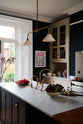 An oblique view of a simple and elegant mushroom-brown Shaker kitchen features a cooker run with freestanding cupboards, a Carrara marble worktop, a large kitchen island with a single Belfast sink, and a small section of copper worktop. Adorned with artisan ceramics, a playful framed illustration, gem lettuce, bright red rhubarb, elegant florals in a vase, and a deVOL Heirloom gaselier, the space feels loving and functional.