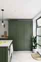 A closer view of the full-height cupboards of a bright, sophisticated Bakehouse Green shaker kitchen, styled with Scandinavian furniture and a calming green palette. The kitchen features a cooker run, a central island with a double Belfast sink and Silestone worktop. Natural light floods the kitchen and dining room through the glass French doors.