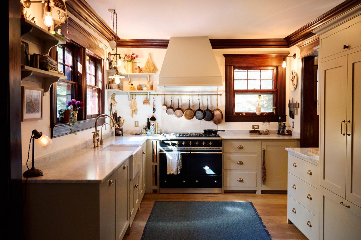 A humble mushroom-painted shaker kitchen features an L-shaped run with a cooker and sink, a shaker canopy, a full-height pantry cabinet, shaker shelves, and aged brass hanging rails for pans. In a small corner of the adjacent hallway is a full-height cabinet with glazed cupboards for storing fruit and an eclectic collection of ceramics.