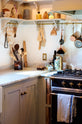A tight and busy corner of a humble mushroom-painted shaker kitchen between the L-shaped cooker and sink run with a Carrara marble worktop. The shaker shelves with aged brass hanging rails hold all sorts of kitchen items, from a triangle bell and a small basket of garlic to measuring spoons, cleaning brushes, and pans.