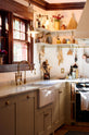 An angled view of a humble mushroom-painted shaker kitchen features an L-shaped cooker and sink run, a shaker canopy, a full-height pantry cabinet, shaker shelves, and aged brass hanging rails for pans. There is a single Belfast sink with an aged brass Ionian tap and wonderfully collected ceramics and kitchen items.