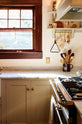 A busy corner of a humble mushroom-painted shaker kitchen between the L-shaped cooker and sink run with a Carrara marble worktop. The shaker shelves with aged brass hanging rails hold all sorts of kitchen items, from a triangle bell and a small basket of garlic to a ceramic mug with a collection of wooden spoons. The rich brown-framed window adds natural light and serenity to the kitchen.