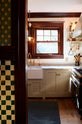 Peeking through a green-and-white mosaic wall and moss-green wallpaper is a humble mushroom-painted shaker kitchen. It features a single Belfast sink with an aged brass Ionian tap and a Carrara marble worktop. A rich brown-framed window floods the space with natural light and serenity.