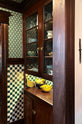 Peeking through a green-and-white mosaic wall and moss-green wallpaper hallway with a full-height cabinet with glazed cupboards for storing fruit and an eclectic collection of ceramics.