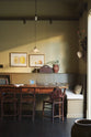 A simple buttery yellow Shaker living space features a Folgate green bespoke bench seat with an open base cabinet, a shelf, a rise and fall pendant light, and a cohesive set of vintage table and chairs. Lovingly adorned with artisan ceramics, paintings, books, a coat rack and cushions, creating a cosy and Mediterranean cafe-inspired social space.