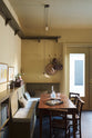 A simple buttery yellow Shaker living space features a Folgate green bespoke bench seat with an open base cabinet, a shelf, a rise and fall pendant light, and a cohesive set of vintage table and chairs. Lovingly adorned with artisan ceramics, paintings, books, a coat rack and cushions, creating a cosy and Mediterranean cafe-inspired social space.