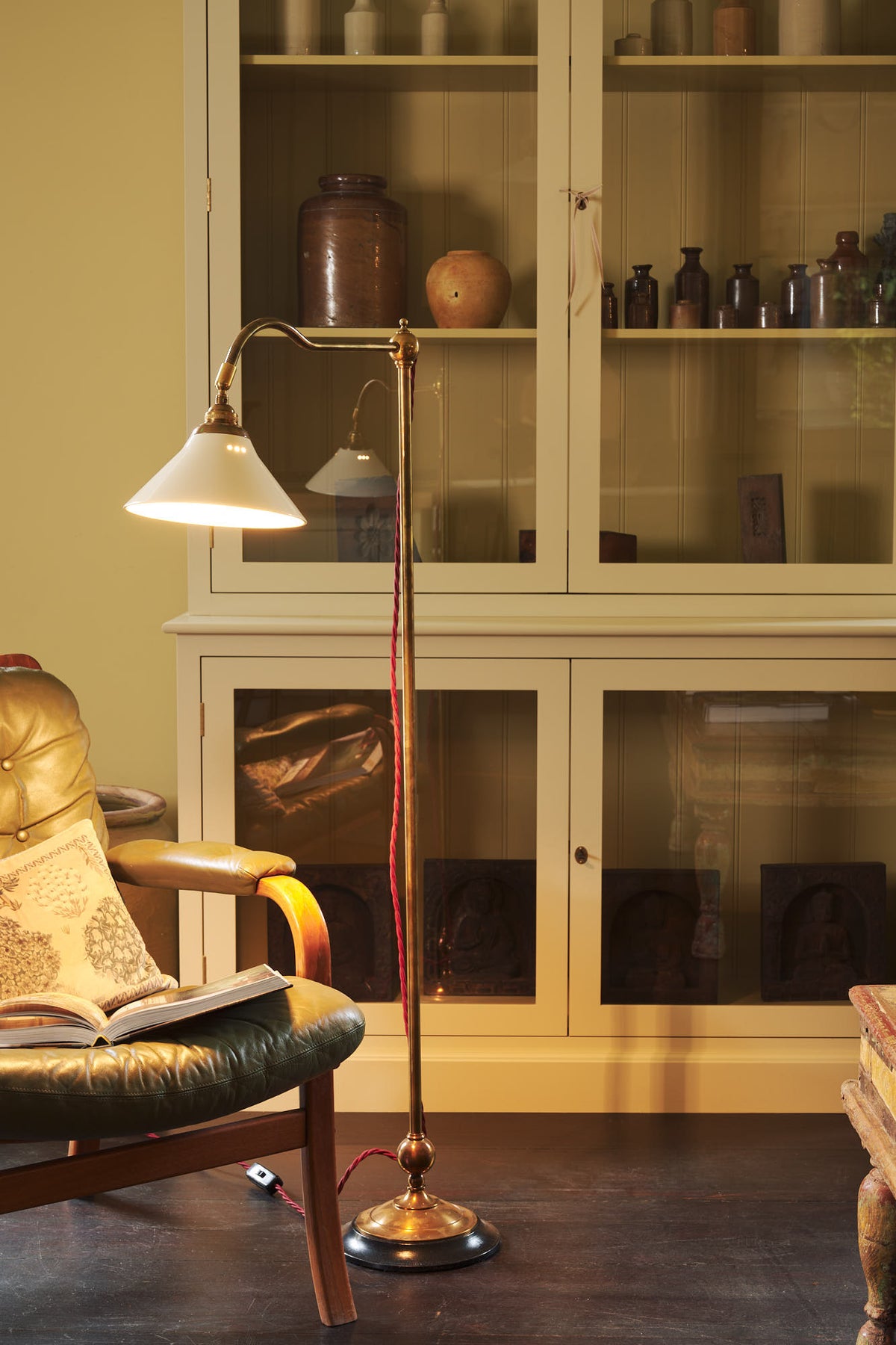 A close-up of a simple buttery yellow Shaker living space features a Quaker cream curiosity cupboard, a grand tour reading lamp, and a cohesive set of vintage furniture. Lovingly adorned with artisan ceramics, books, and antique canes, creating a humble and mid-century inspired social space