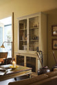 An oblique view of a simple buttery yellow Shaker living space features a Quaker cream curiosity cupboard, a grand tour reading lamp, and a cohesive set of vintage furniture. Lovingly adorned with artisan ceramics, books, and antique canes, creating a humble and mid-century inspired social space.