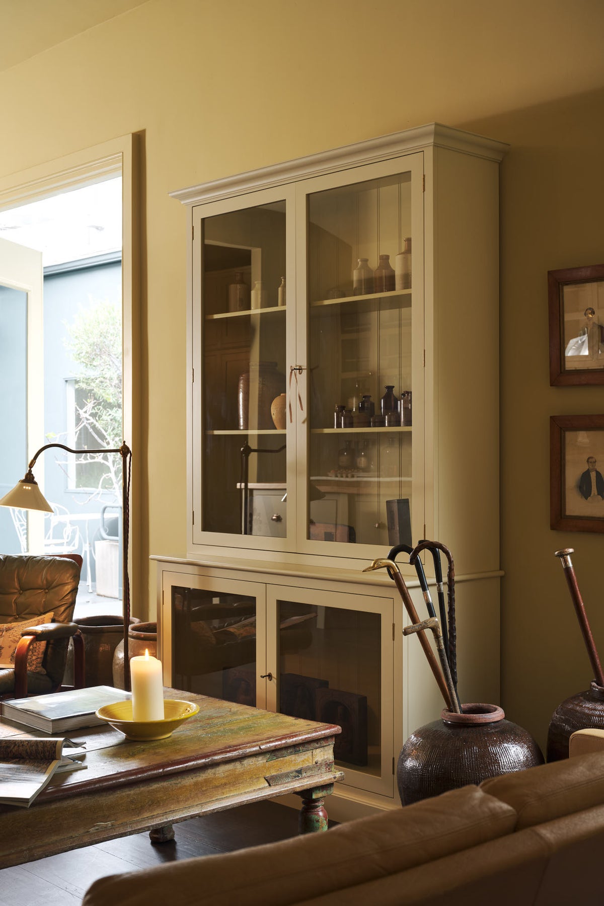 An oblique view of a simple buttery yellow Shaker living space features a Quaker cream curiosity cupboard, a grand tour reading lamp, and a cohesive set of vintage furniture. Lovingly adorned with artisan ceramics, books, and antique canes, creating a humble and mid-century inspired social space.