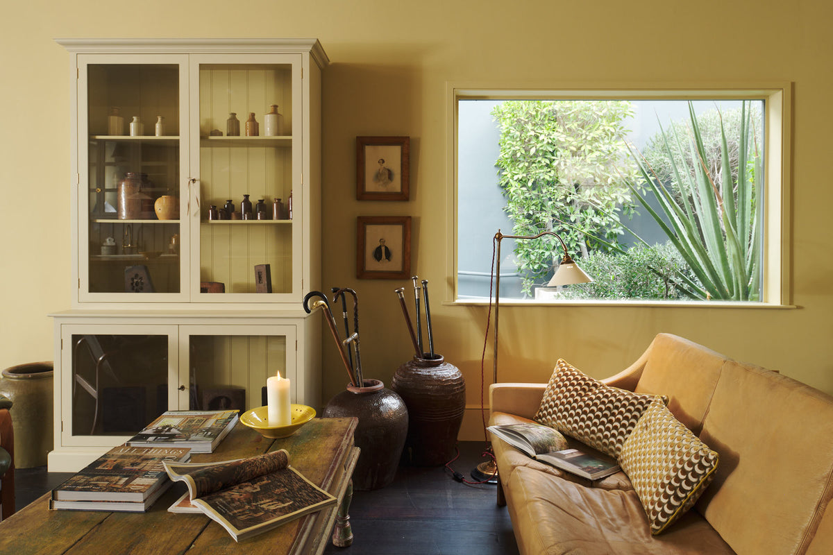 A simple buttery yellow Shaker living space features a Quaker cream curiosity cupboard, a grand tour reading lamp, and a cohesive set of vintage furniture. Lovingly adorned with artisan ceramics, books, and antique canes, creating a humble and mid-century inspired social space.