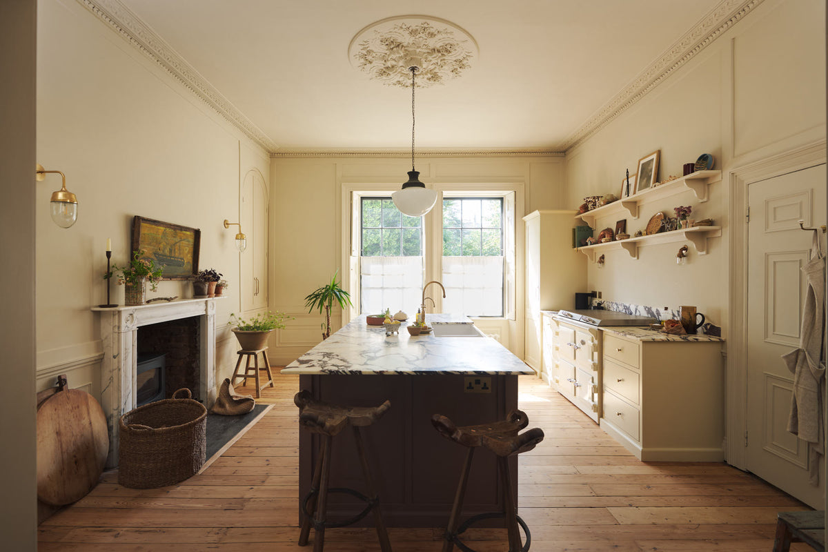 A bright, spacious, warm, and simple-yet-grand shaker kitchen featuring a large kitchen island with a Calacatta Viola worktop, a cooker run, a fridge/freezer cupboard, and long shaker shelves. Perfectly decorated with beloved vintage furniture and decor made from different natural materials, it creates a scene of absolute beauty, personality and functionality.