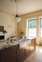 An oblique view of a bright, spacious, warm, and simple-yet-grand shaker kitchen featuring a large kitchen island with a Calacatta Viola worktop, and an aged brass Ionian tap. Perfectly decorated with beloved vintage furniture and decor made from different natural materials, it creates a scene of absolute beauty, personality and functionality.