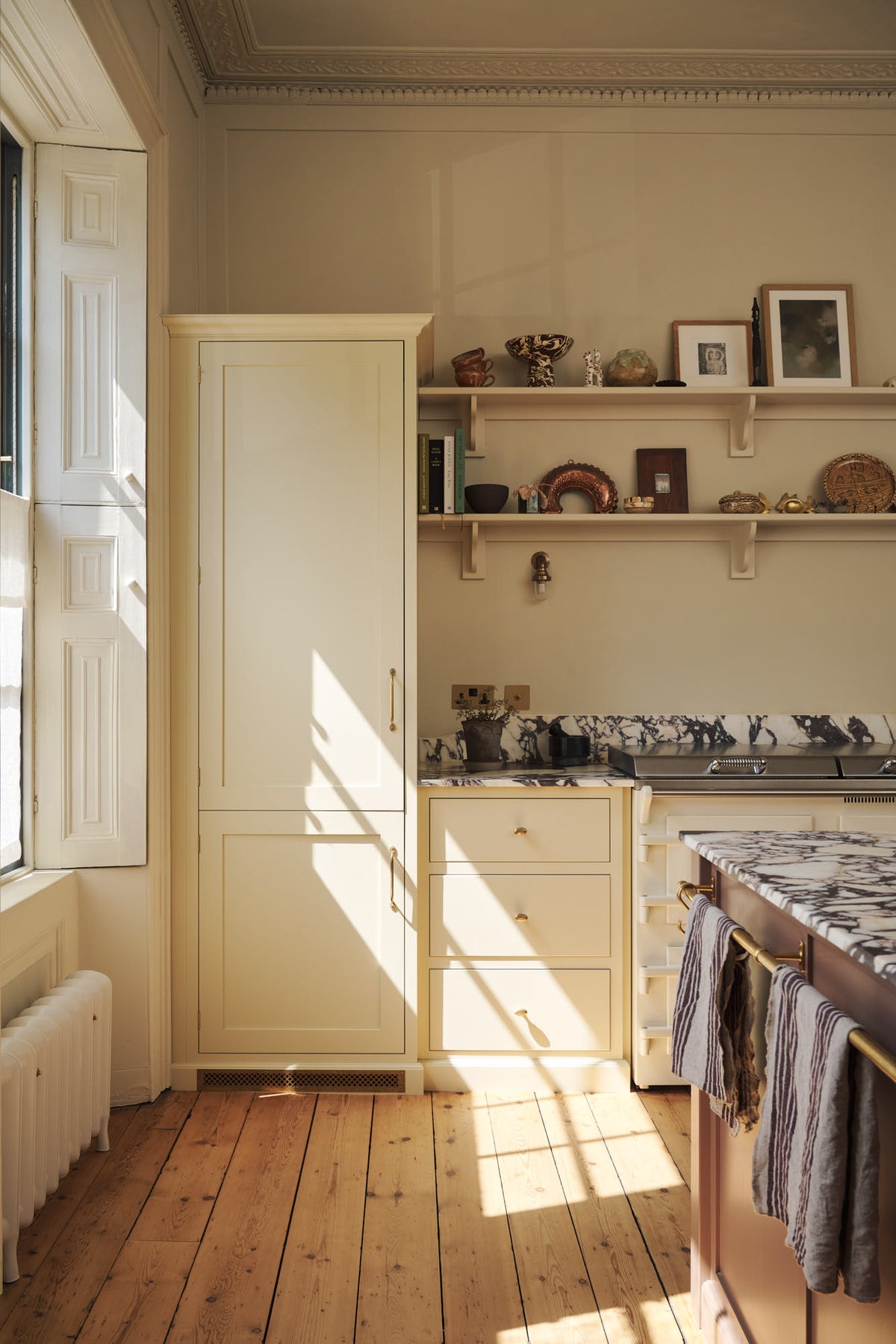 A quiet corner of a bright, spacious, warm, and simple-yet-grand shaker kitchen featuring a large kitchen island with a Calacatta Viola worktop, a cooker run, a fridge/freezer cupboard, and long shaker shelves. Perfectly decorated with beloved vintage furniture and decor made from different natural materials, it creates a scene of absolute beauty, personality and functionality.