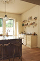 An oblique view of a bright, spacious, warm, and simple-yet-grand shaker kitchen featuring a large kitchen island with a Calacatta Viola worktop, a cooker run, a fridge/freezer cupboard, and long shaker shelves. Perfectly decorated with beloved vintage furniture and decor made from different natural materials, it creates a scene of absolute beauty, personality and functionality.