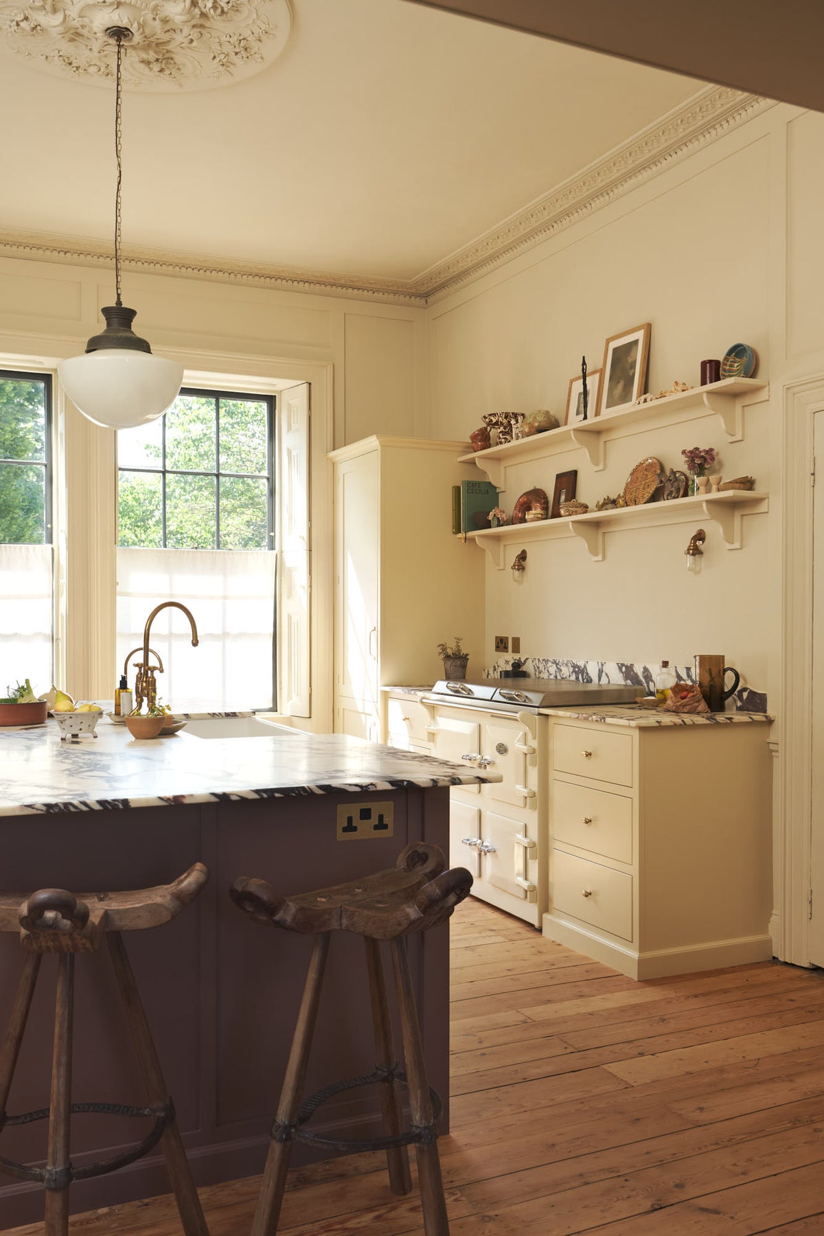 An oblique view of a bright, spacious, warm, and simple-yet-grand shaker kitchen featuring a large kitchen island with a Calacatta Viola worktop, a cooker run, a fridge/freezer cupboard, and long shaker shelves. Perfectly decorated with beloved vintage furniture and decor made from different natural materials, it creates a scene of absolute beauty, personality and functionality.