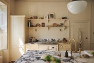 A bright, spacious, warm, and simple-yet-grand shaker kitchen featuring a large kitchen island with a Calacatta Viola worktop, a cooker run, a fridge/freezer cupboard, and long shaker shelves. Perfectly decorated with beloved vintage furniture and decor made from different natural materials, it creates a scene of absolute beauty, personality and functionality.