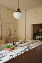An oblique view of a bright, spacious, warm, and simple-yet-grand shaker kitchen featuring a large kitchen island with a Calacatta Viola worktop, a double sink, and an aged brass Ionian tap. Perfectly decorated with beloved vintage furniture and decor made from different natural materials, it creates a scene of absolute beauty, personality and functionality.