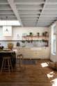 A spacious, Mediterranean-inspired Quaker Cream Shaker kitchen features a long sink and cooker run with a Carrara marble worktop, and a classic oak shelf. Lovingly decorated with a wall of vibrant blue patterned splashback tiles, aged brass hanging rails, Helen’s Stools, green house plants, and fresh produce, it creates a wonderfully social and practical kitchen space.