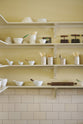 A close-up of a simple Quaker cream and Folgate green Shaker scullery features architectural glazed partitions, and bespoke shelving, finished with a cream subway tiles splashback. Lovingly adorned with artisan ceramics, wooden chopping boards, and antique kitchen accessories, it gives a sombre and practical space.