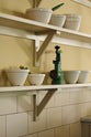 A close-up of a simple Quaker cream and Folgate green Shaker scullery features architectural glazed partitions, and bespoke shelving, and a sink run with a Silestone worktop, finished with a cream subway tiles splashback. Lovingly adorned with artisan ceramics, and antique kitchen accessories, it gives a sombre and practical space.