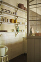 An oblique view of a simple Quaker cream and Folgate green Shaker scullery features architectural glazed partitions, bespoke shelving, and a sink run with a Silestone worktop, finished with a cream subway tiles splashback. Lovingly adorned with artisan ceramics, wooden chopping boards, and antique kitchen accessories and furniture, it gives a sombre and practical space.