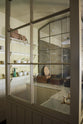 An oblique view of a simple Quaker cream and Folgate green Shaker scullery features architectural glazed partitions, bespoke shelving, and a sink run with a Silestone worktop, finished with a cream subway tiles splashback. Lovingly adorned with artisan ceramics, wooden chopping boards, and antique kitchen accessories and furniture, it gives a sombre and practical space.