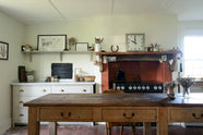 A small and quaint mushroom-coloured Shaker kitchen features a sink run made up of a mix of stave hardwood and Carrara marble worktops, a shelf, and a black stove nestled inside the original building’s chimney breast. Beautifully decorated with vintage furniture such as a large solid oak table, a silver scale, and assorted bric-a-brac, it creates a lovely social space full of character.