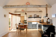 A small and humble pantry-blue Shaker kitchen features an L-shaped sink and cooker run with a stave hardwood worktop, a single Belfast sink, and aged brass Mayan taps. Beautifully decorated with a taxidermy bird sitting on the roof’s wooden beams, vintage furniture, books, and assorted bric-a-brac, creates a heartwarming family farm kitchen space.