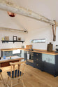 An oblique view of a small and humble pantry-blue Shaker kitchen features an L-shaped sink and cooker run with a stave hardwood worktop, a single Belfast sink, and aged brass Mayan taps. Beautifully decorated with a livestock bell attached on the roof’s wooden beams, vintage furniture, framed still-life paintings, and assorted bric-a-brac, creates a heartwarming family farm kitchen space.