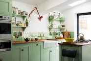 An oblique view of a vibrant pea-green Shaker kitchen features a sink run and a kitchen island with a gas cooker, both topped with a reclaimed iroko school countertop, matching green square splashback tiles, shelves with brass hanging rails, and a double oven cabinet with drawers. Beautifully decorated with artisan ceramics, dried ingredients in a glass jar, oranges, and a clamped red lamp, creates a friendly and welcoming kitchen.