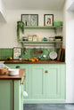 A corner of a pea-green Shaker kitchen features a sink run and a kitchen island with a gas cooker, both topped with a reclaimed iroko school countertop, matching green square splashback tiles, and shelves with brass hanging rails. Beautifully decorated with artisan ceramics, lemons, oranges, and a string-of-pearls plant, it creates a friendly and welcoming kitchen.