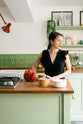 A corner of a pea-green Shaker kitchen with Rachel Khoo features a sink run and a kitchen island with a gas cooker, both topped with a reclaimed iroko school countertop, matching green square splashback tiles, shelves with brass hanging rails, and a single Belfast sink with aged brass Mayan taps. Beautifully decorated with artisan ceramics, lemons, oranges, and a string-of-pearls plant, it encourages a friendly and welcoming kitchen space.
