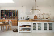 A minimalist Damask off-white shaker kitchen with a solid oak island and Carrara marble worktops for the sink and cooker. The sink and cooker runs feature crackle metro splashback tiles. A shaker table with oak chairs and glazed countertop cupboards complete the space, accented by warm browns and golds, from wooden chopping boards to the aged brass Heirloom gaselier light.
