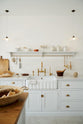 A closer look at a Damask off-white shaker kitchen featuring a solid oak island and Carrara marble worktops on the sink and cooker runs. The sink run includes a single fluted Belfast sink with an aged brass Ionian tap. Crackle metro splashback tiles adorn the walls, while a Shaker shelf with aged brass rails holds essentials like scissors, scrubs, and a grater.