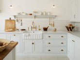 A closer look at a Damask off-white shaker kitchen featuring a solid oak island and Carrara marble worktops on the sink and cooker runs. The sink run includes a single fluted Belfast sink with an aged brass Ionian tap. Crackle metro splashback tiles adorn the walls, while a Shaker shelf with aged brass rails holds essentials like scissors, scrubs, and a grater.
