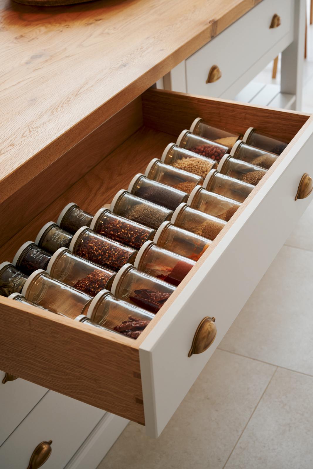A closer look at the drawer of a solid oak island in a Damask off-white Shaker kitchen, neatly lined with clear jars of dried ingredients, herbs, and spices.