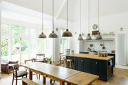 An angled view of a high-ceiling, pantry blue shaker kitchen features a sink and cooker run with a Silestone worktop, complemented by a large kitchen island topped with solid oak and glazed cupboards. The cooker run sits in an alcove with white splashback tiles, topped by a shaker shelf displaying assorted ceramic and wooden bric-a-bracs. Rusty metal hanging lights in the centre add an industrial touch to the space.