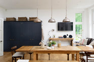 Showing the dinning area of the pantry blue kitchen, there’s two full-height pantry cupboards, a flat screen tv, a vintage oak dining table with wooden benches and mismatched chairs adds character, while rusty metal pendant lights in the centre bring an industrial edge.