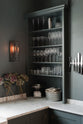 A quiet corner of a charcoal-black, classic shaker kitchen features a U-shaped sink and cooker run with Carrara marble worktop, a double Belfast sink, chrome Ionian tap and open countertop cupboards. Beautifully decorated with a magnetic kitchen knife holder, a steampunk wall light, a steel weight, branches of flowers, and the original Georgian wooden sliders, the family kitchen strikes a wonderful balance between modern style and timeless character.