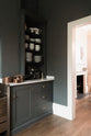 A quiet corner of a charcoal-black classic Shaker kitchen features a U-shaped sink and cooker run with a Carrara marble worktop, matching splashback, and open countertop cupboards. Beautifully decorated with copper pans, simple white ceramic plates and bowls, and a black espresso maker, the family kitchen strikes a wonderful balance between modern style and timeless character.