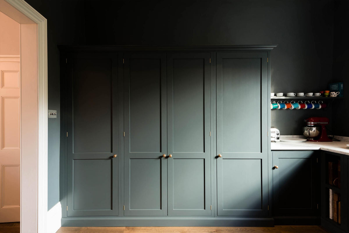 A charcoal-black, classic shaker kitchen features a U-shaped sink and cooker run with Carrara marble worktop and a bank of fitted pantry cabinets. Beautifully decorated with a colourful collection of mugs, a striking red mixer, and a silver toaster machine, the family kitchen strikes a wonderful balance between modern style and timeless character.