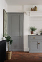 A closer look at the shaker kitchen’s full-height pantry door and the edge of a simple, modern cooker run, finished with white porcelain splashback tiles. Cupboards in mid-grey, terracotta tile flooring, and crisp white walls. Styled with a woven basket plant, a framed vintage illustration, and handcrafted ceramic bowls, jugs, and mugs.