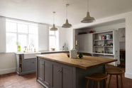 An angled view of a simple, modern shaker kitchen in a historic Cheshire townhouse, featuring a Silestone sink run, a pantry cupboard with open shelves, and a large island with a solid oak worktop and a breakfast nook. Painted in muted cool grey with white walls and warm terracotta tile flooring.