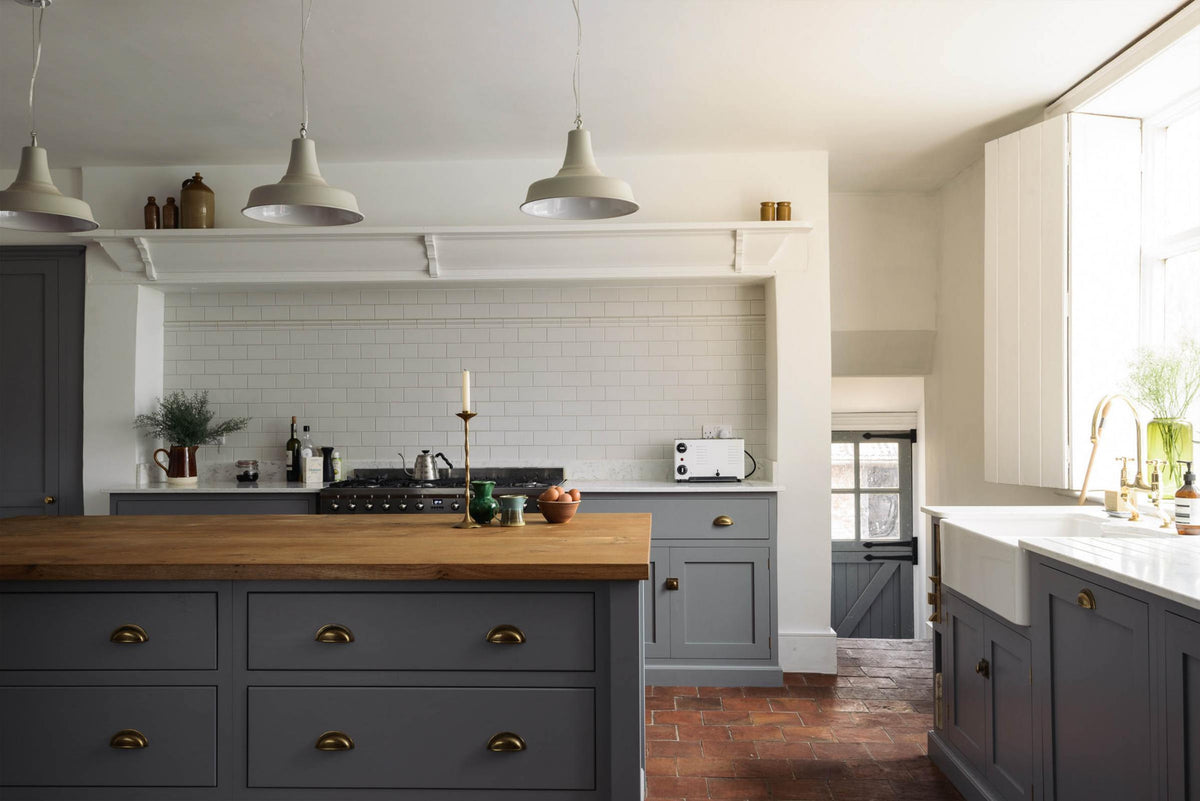 A closer look at a simple, modern shaker kitchen in a historic Cheshire townhouse, featuring a cooker run, Silestone sink run with a Belfast sink, aged brass Ionian tap, and a large island with a solid oak worktop and breakfast nook. Painted in muted cool grey with white walls and warm terracotta tile flooring.