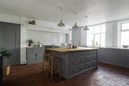 An angled view of a simple, modern shaker kitchen in a historic Cheshire townhouse, featuring a Silestone sink run with a Belfast sink and aged brass Ionian tap, and a large island with a solid oak worktop and a breakfast nook. Painted in muted cool grey with white walls and warm terracotta tile flooring.