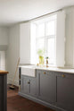 A close and angled view at a simple, modern shaker kitchen in a historic Cheshire townhouse, featuring a cooker run, Silestone sink run with a Belfast sink, aged brass Ionian tap, and a large island with a solid oak worktop and breakfast nook. Painted in muted cool grey with white walls and warm terracotta tile flooring.